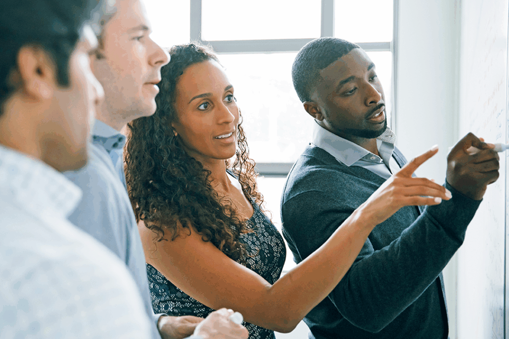 Four professionals in a brightly lit office collaborate, looking focused while pointing and writing notes on a whiteboard.