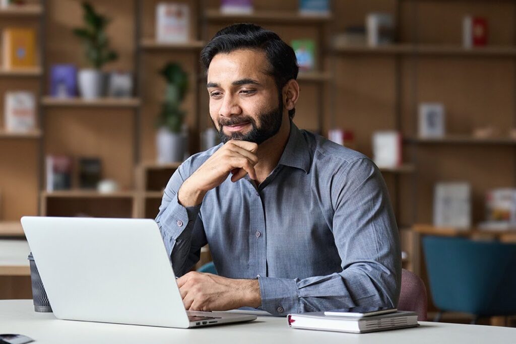 A smiling man in a well-lit workspace works at a laptop; notebooks and a coffee cup sit beside the computer.