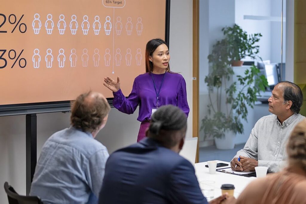 A woman in a purple blouse presents diversity statistics on a screen to colleagues seated around a conference table in a modern office meeting room.