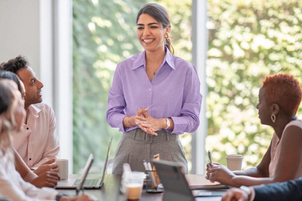 A smiling woman in a purple shirt leads a meeting with colleagues.